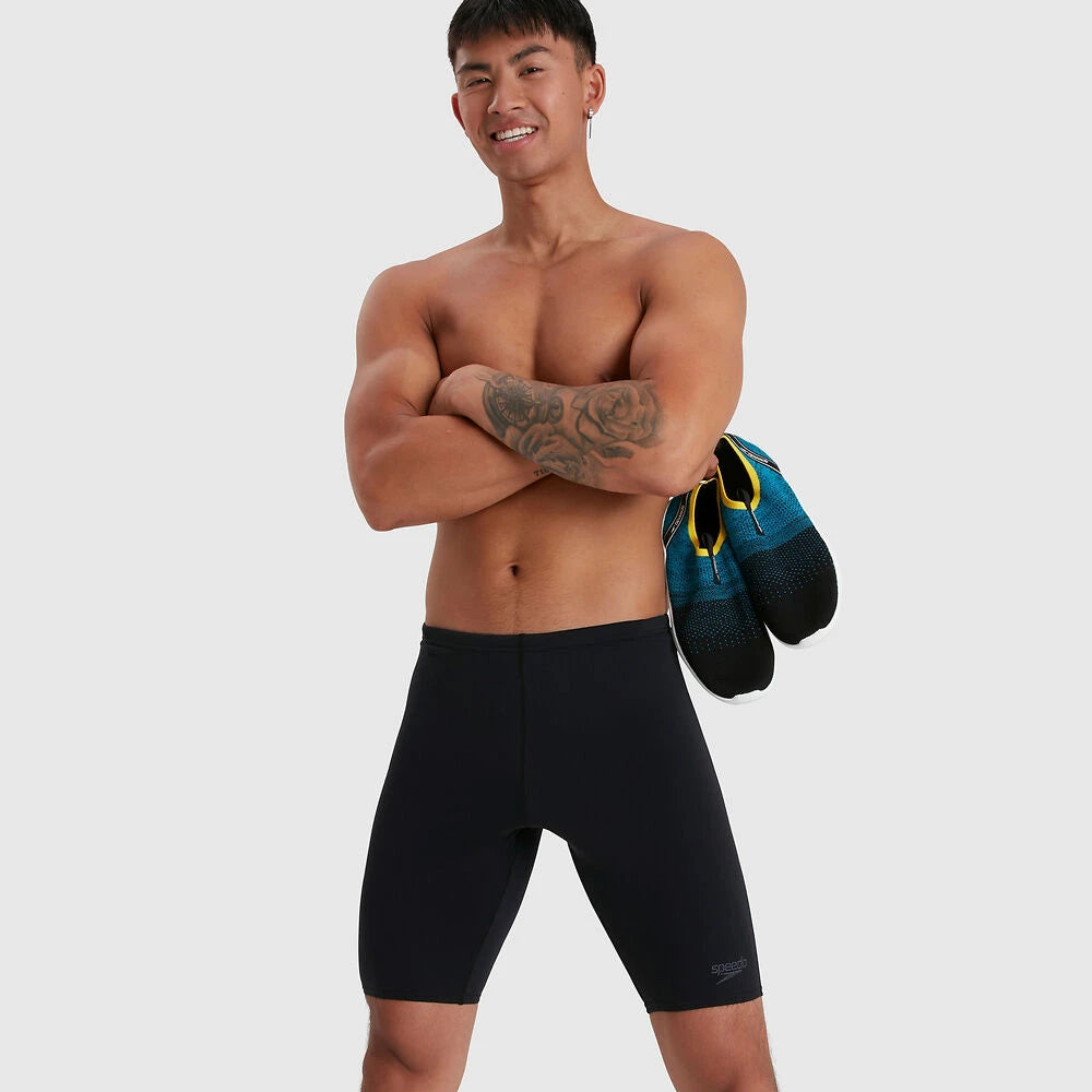 A man standing and posing with shoes in his hand, wearing black Speedo jammers.