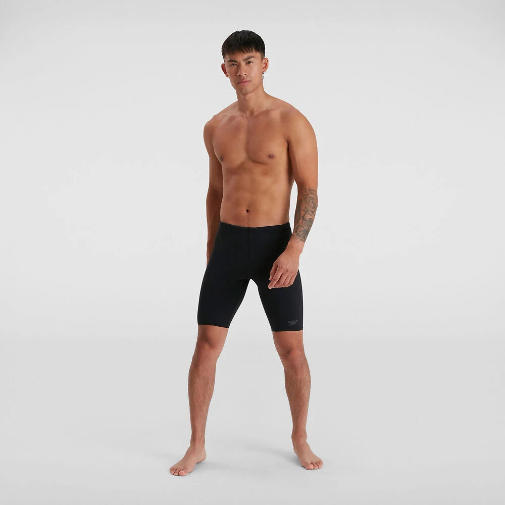 A man standing and posing with shoes in his hand, wearing black Speedo jammers.