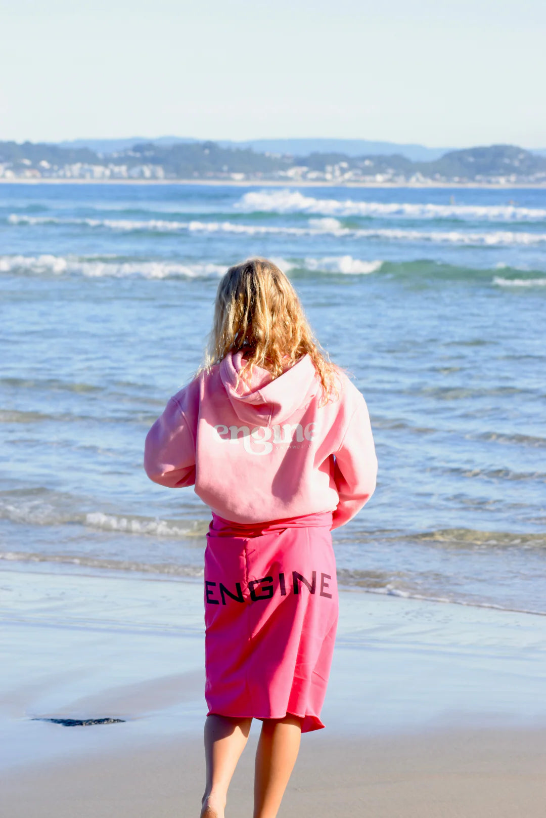Woman on a beach holding a pink towel with 'Engine' branding