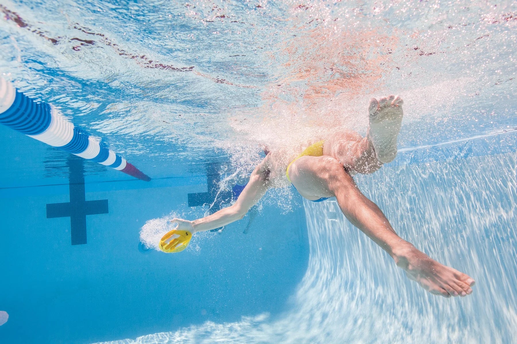 Person swimming underwater in a pool using Finis yellow hand paddles