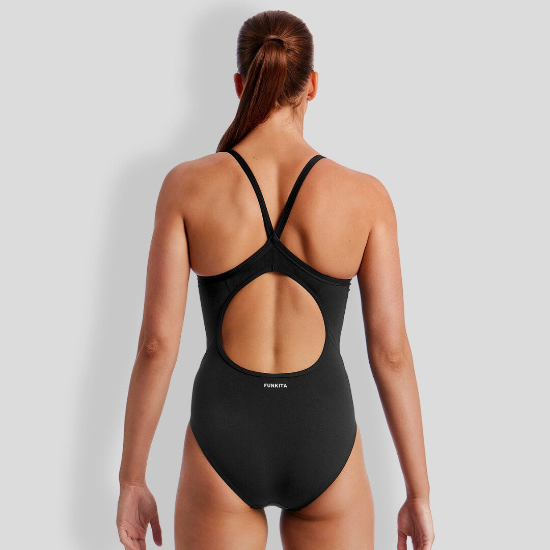 A woman viewed from the back wearing a black one-piece swimsuit with a open-back cutout and thin crisscross straps,
