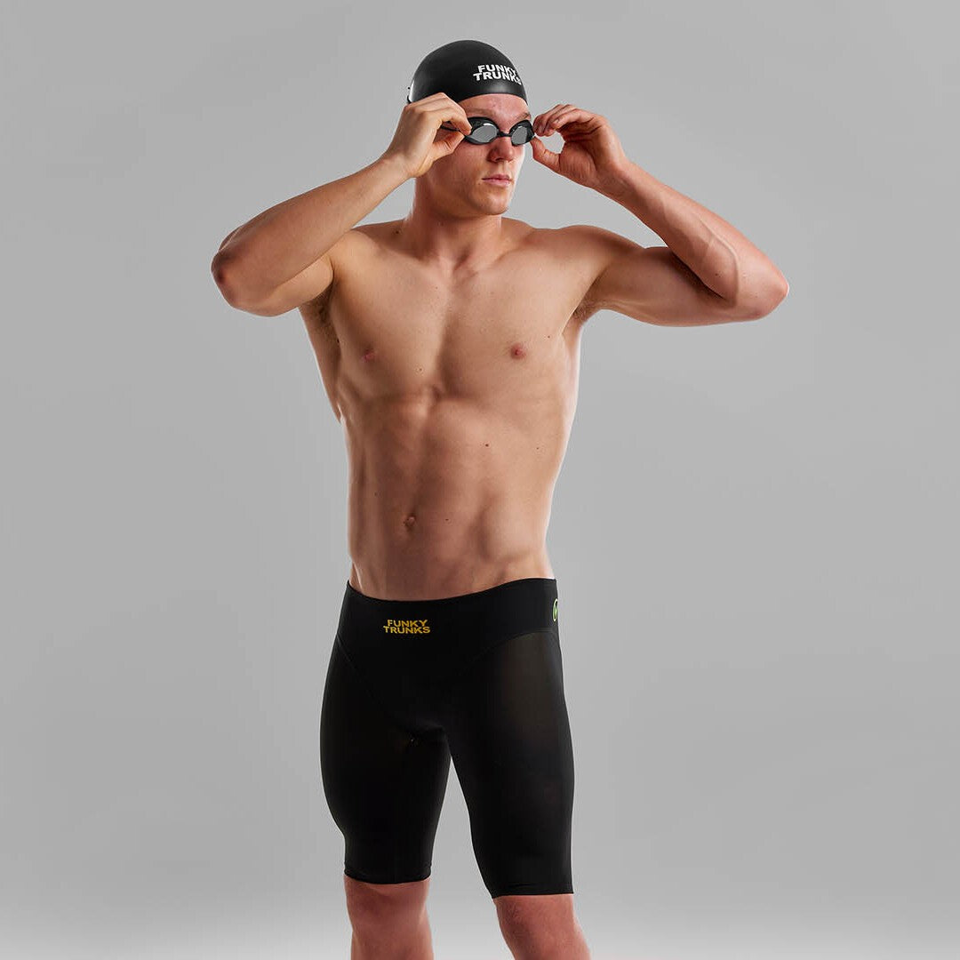 A man wearing Funky Trunks Black Attack Men's Apex Viper Jammers with a black Funky Trunks swim cap, adjusting his goggles.