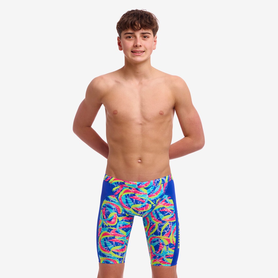A boy with his hands behind his back wearing multicolored Funky Trunks Choppy Waters training jammers with the Funky Trunks brand logo on the leg.