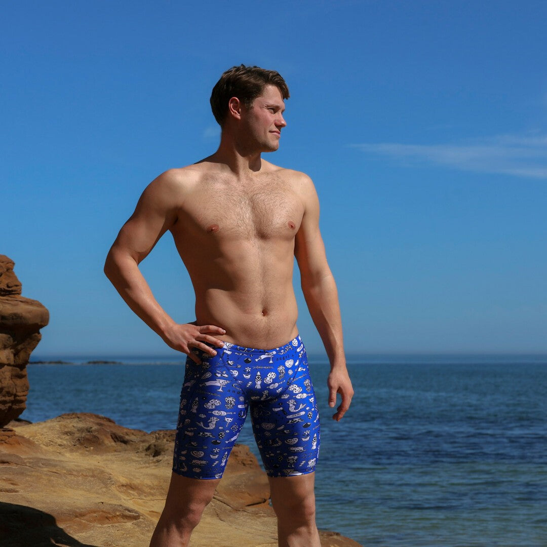 A shirtless man standing on a rocky beach wearing blue swimming jammers with a white "scratchy" pattern from Funky Trunks with the ocean and blue sky in the background.