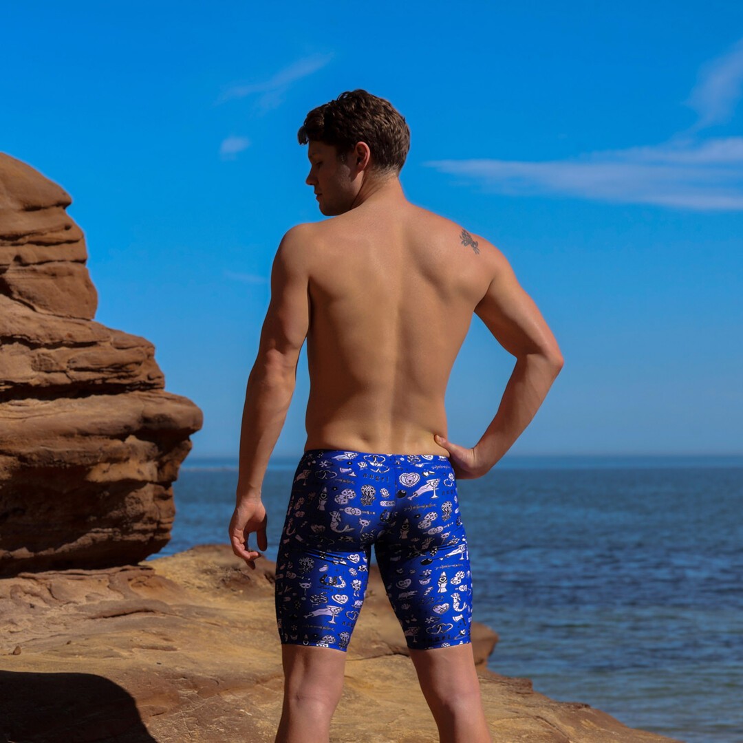 A shirtless man with his back to the camera standing on a rocky beach wearing blue swimming jammers with a white "scratchy" pattern from Funky Trunks with the ocean and blue sky in the background.