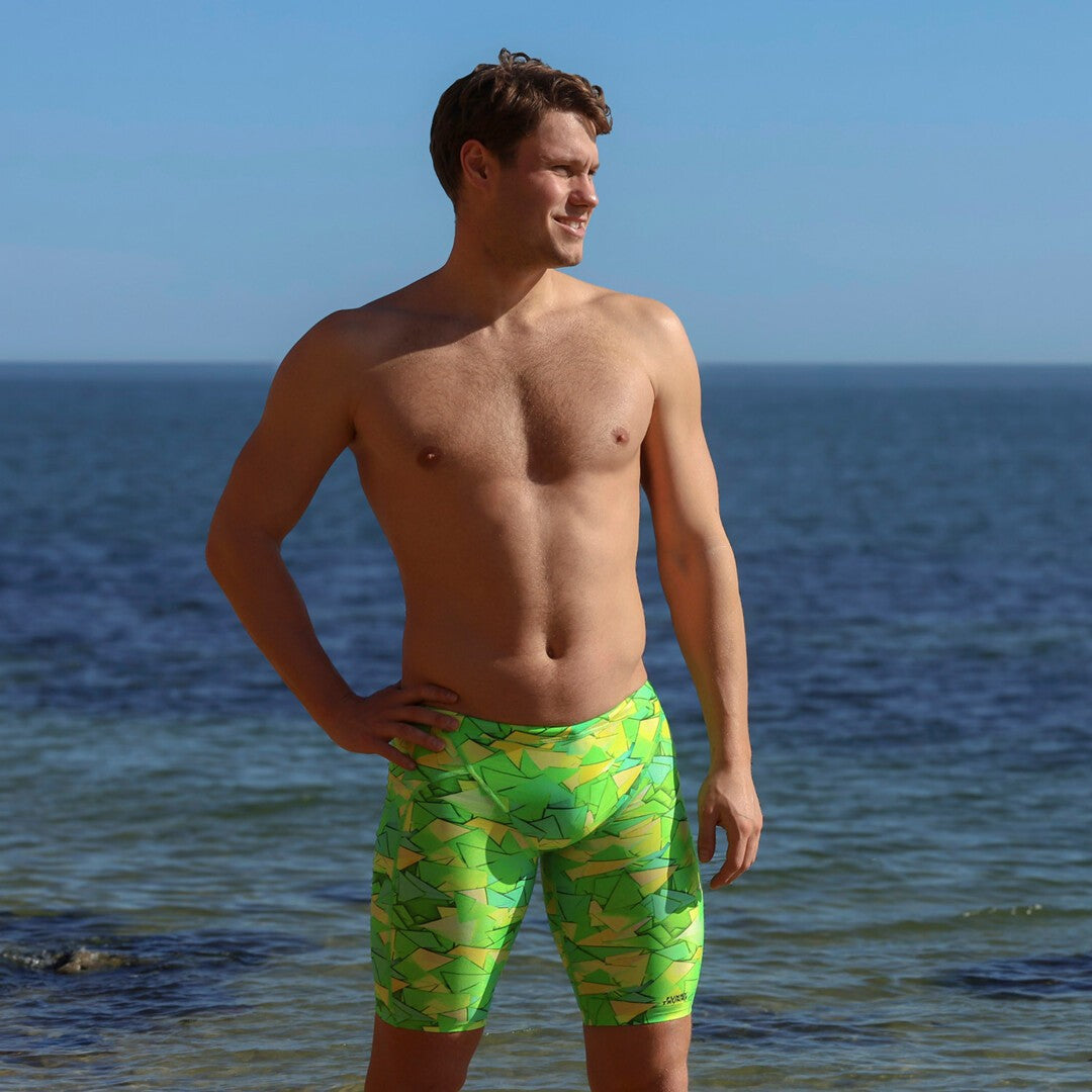 A man standing at the beach wearing bright green and yellow patterned swimming jammers from Funky Trunks, with the ocean and blue sky in the background.