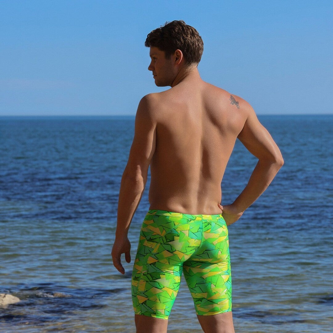 A man standing at the beach with his back to the camera wearing bright green and yellow patterned swimming jammers from Funky Trunks, with the ocean and blue sky in the background.