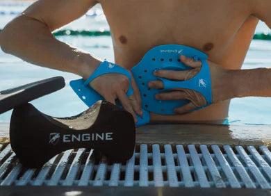 Person wearing blue Engine hand paddles with a black Engine branded pullbuoy on a pool deck.