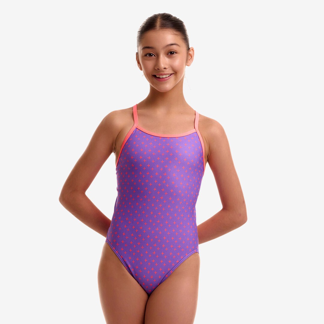 A young girl modeling a purple and pink patterned one-piece swimsuit against a light gray studio background from the brand Funkita.