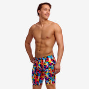 Man wearing colourful geometric swim jammers with a multicolor pattern of blue, red, yellow, and pink triangles, against a white studio background.