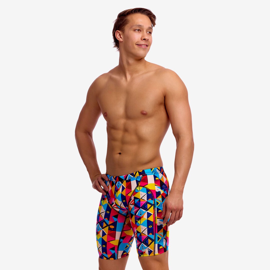 Man wearing colourful geometric swim jammers with a multicolor pattern of blue, red, yellow, and pink triangles, against a white studio background.