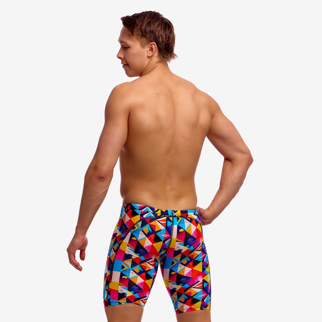Back view of a man wearing colourful geometric swim jammers with a multicolor pattern of blue, red, yellow, and pink triangles, against a white studio background.
