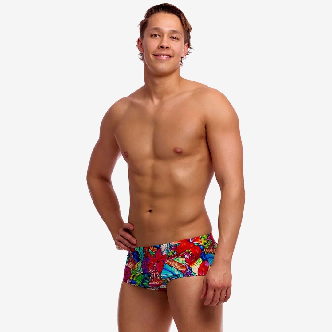 A shirtless man with one hand on his hip wearing bright multicoloured floral print swim trunks