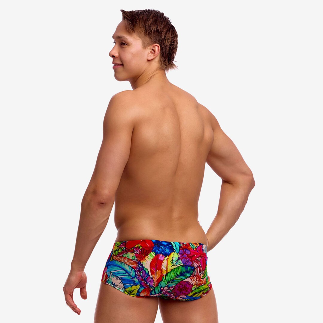 A shirtless man from behind with one hand on his hip wearing bright multicoloured floral print swim trunks