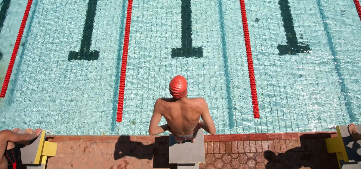 Swimmer with red cap poised at starting block near pool edge, lanes marked by red dividers and numbers visible underwater.
