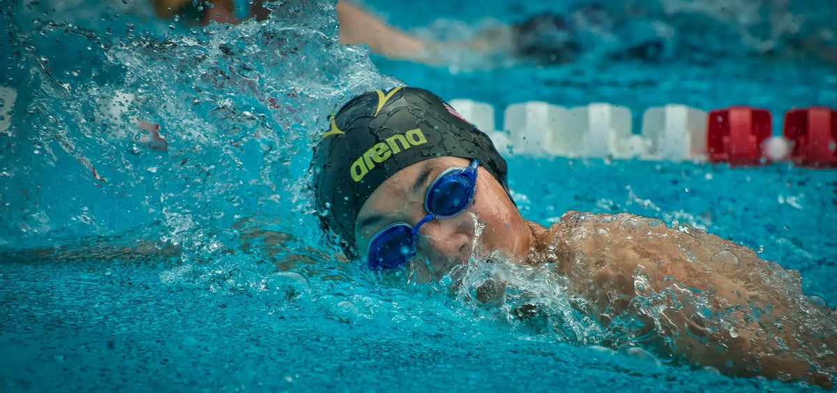 Swimmer wearing an Arena cap and blue goggles in action, with water splashing around in a pool during a race.